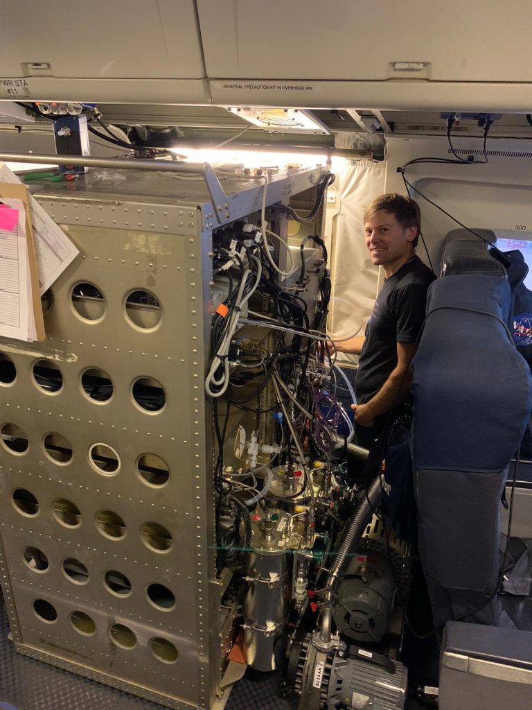 NOAA scientist, Dr. Drew Collins, stands next to a large aerosol measuring appliance with complicated pipes and plumbing housed in the interior of a DC8 plane.