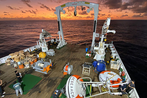 Image of a research vessel with research equipment and a NOAA research buoy on the deck with a colorful sunset in the background. 