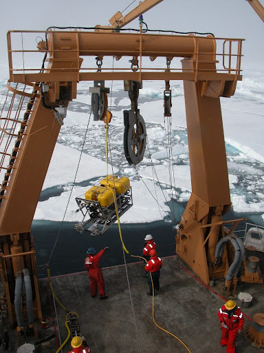 Research vessel navigating through ice in the Arctic Ocean. Crew members are seen on the deck as they deploy a remotely operated vehicle that explores under ice sheets. 