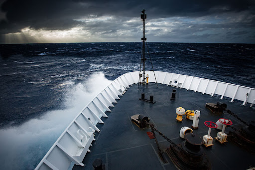 Image of choppy seas and cloudy skies in front of the bow of a large ship.