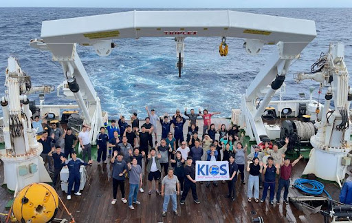 NOAA and KIOS scientists aboard a research cruise in the Pacific Ocean, holding a "KIOS" banner at the stern of the ship.