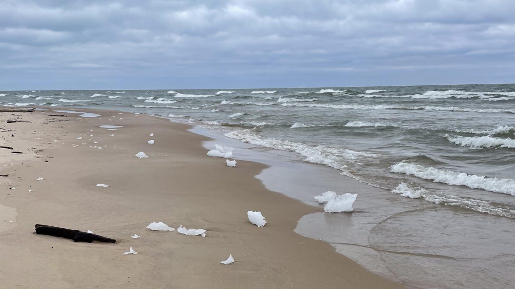 A sandy beach is dotted with chunks of ice. The waves are hitting the beach and there is no ice out on the water.