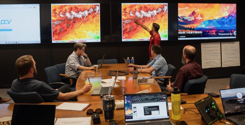 A forecaster points to a large monitor on a wall depicting high winds during a meeting as several colleagues watch from a nearby conference table.
