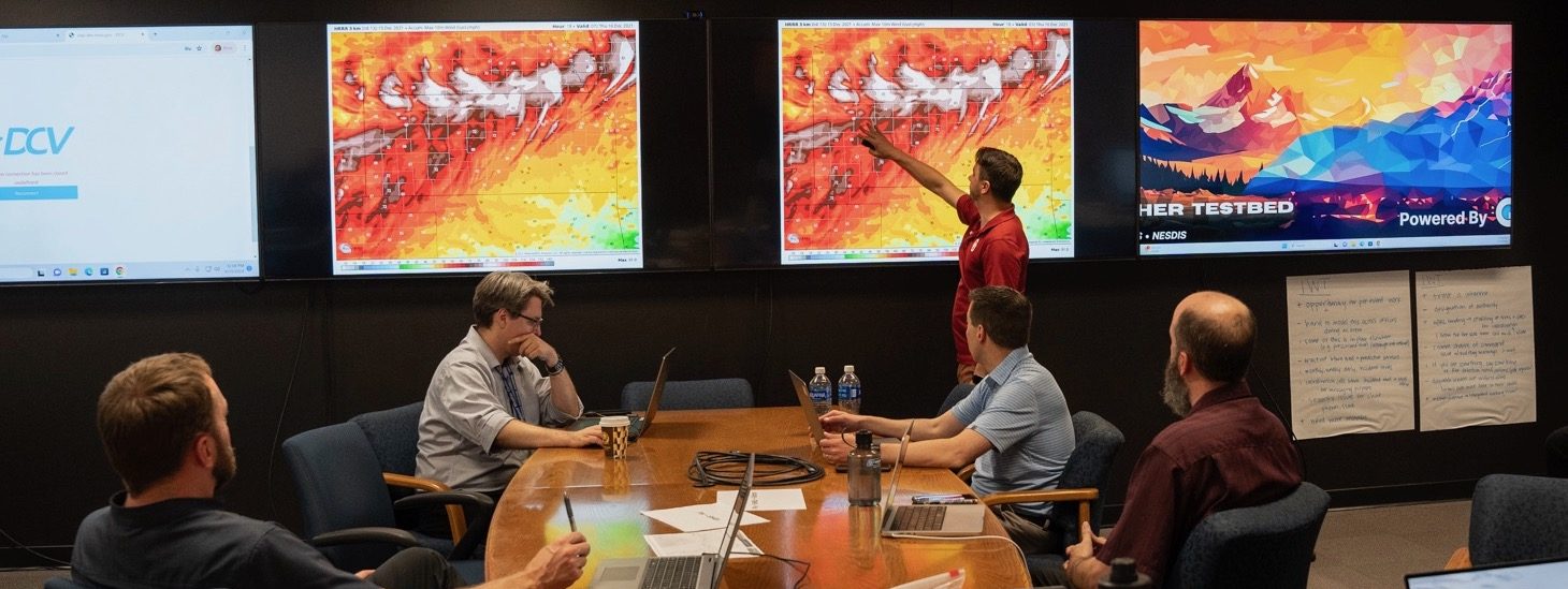 A forecaster points to a large monitor on a wall depicting high winds during a meeting as several colleagues watch from a nearby conference table.