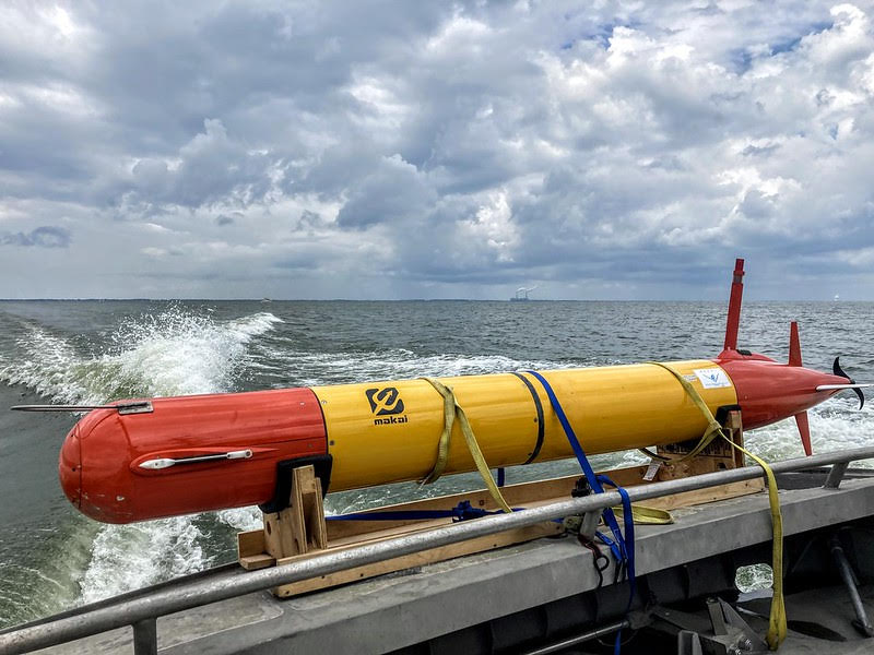 A long, torpedo-shaped device is strapped to the side of a boat that is moving fast through lake water. 